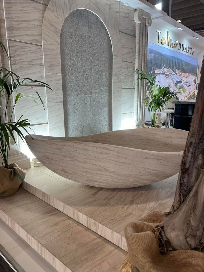Panchina curva in legno bianco in una hall interna illuminata da una luce soffusa, con piante e gradini in pietra.