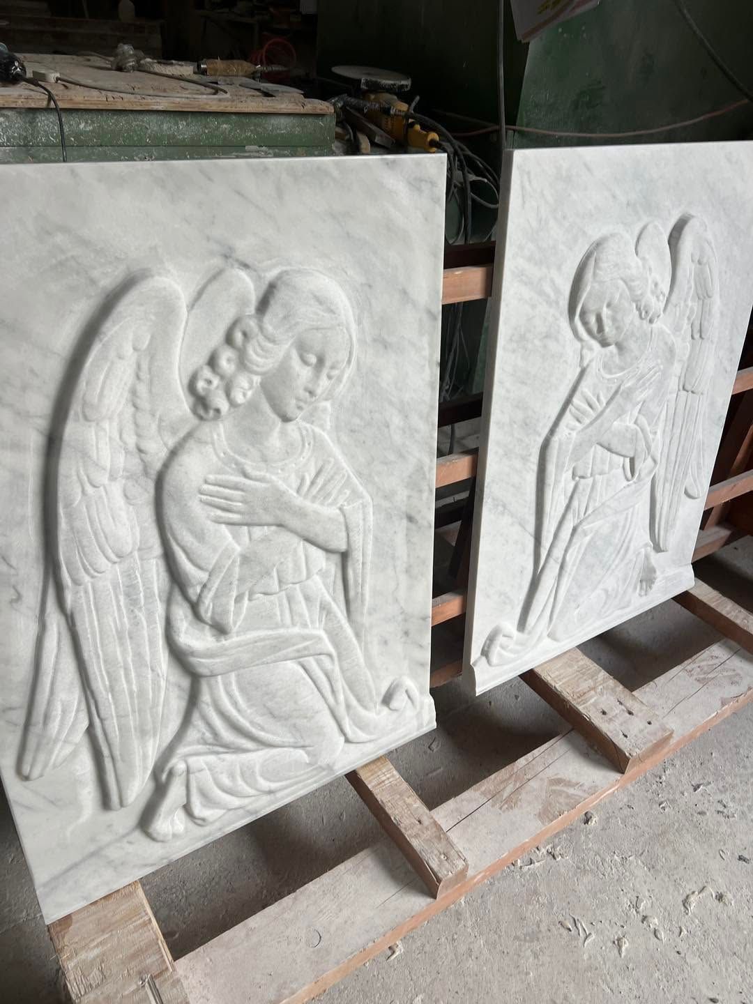 In un'officina, due pannelli in pietra scolpita con bassorilievi di angeli, appoggiati su supporti di legno.