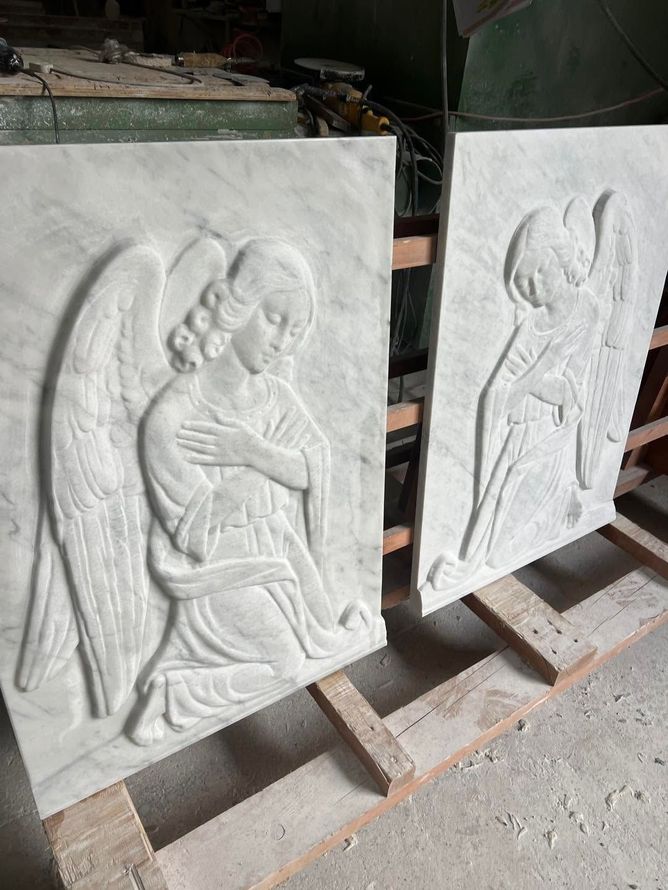 In un'officina, due pannelli in pietra scolpita con bassorilievi di angeli, appoggiati su supporti di legno.