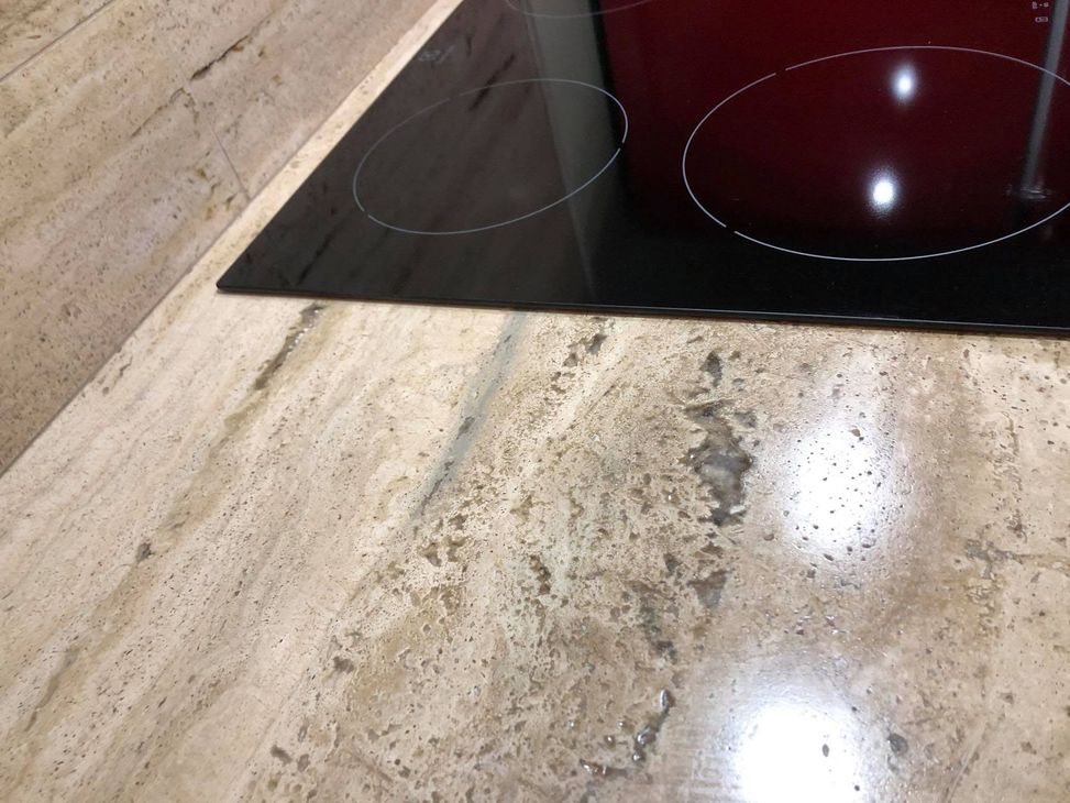 Piano di lavoro della cucina accanto a un piano cottura in vetro nero con luci bianche che si riflettono su una superficie in pietra beige.