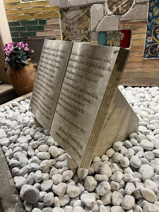 Scultura a forma di libro aperto in metallo, posizionata su ciottoli bianchi in un'ambientazione decorativa da giardino.