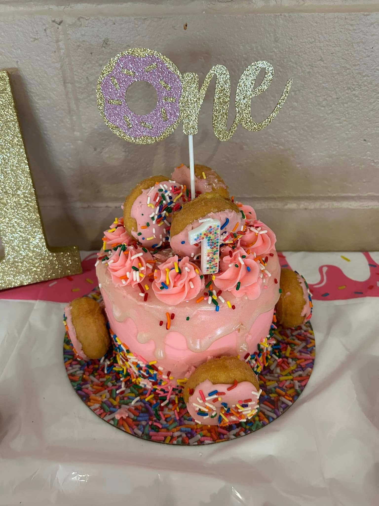 Pink sprinkled cake with gold “one” topper and candles on a decorated table.