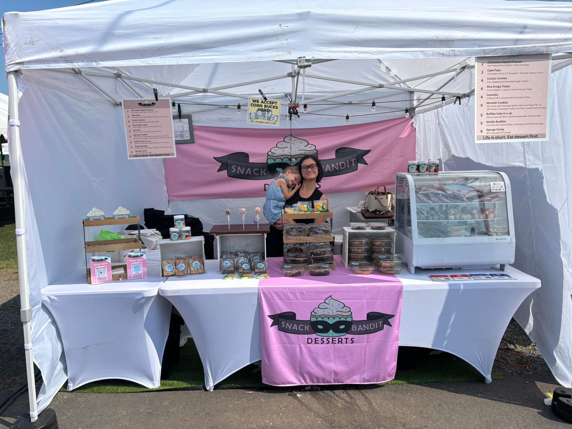 Outdoor vendor booth with pink banners, two people behind a table of baked goods and jars under a white canopy