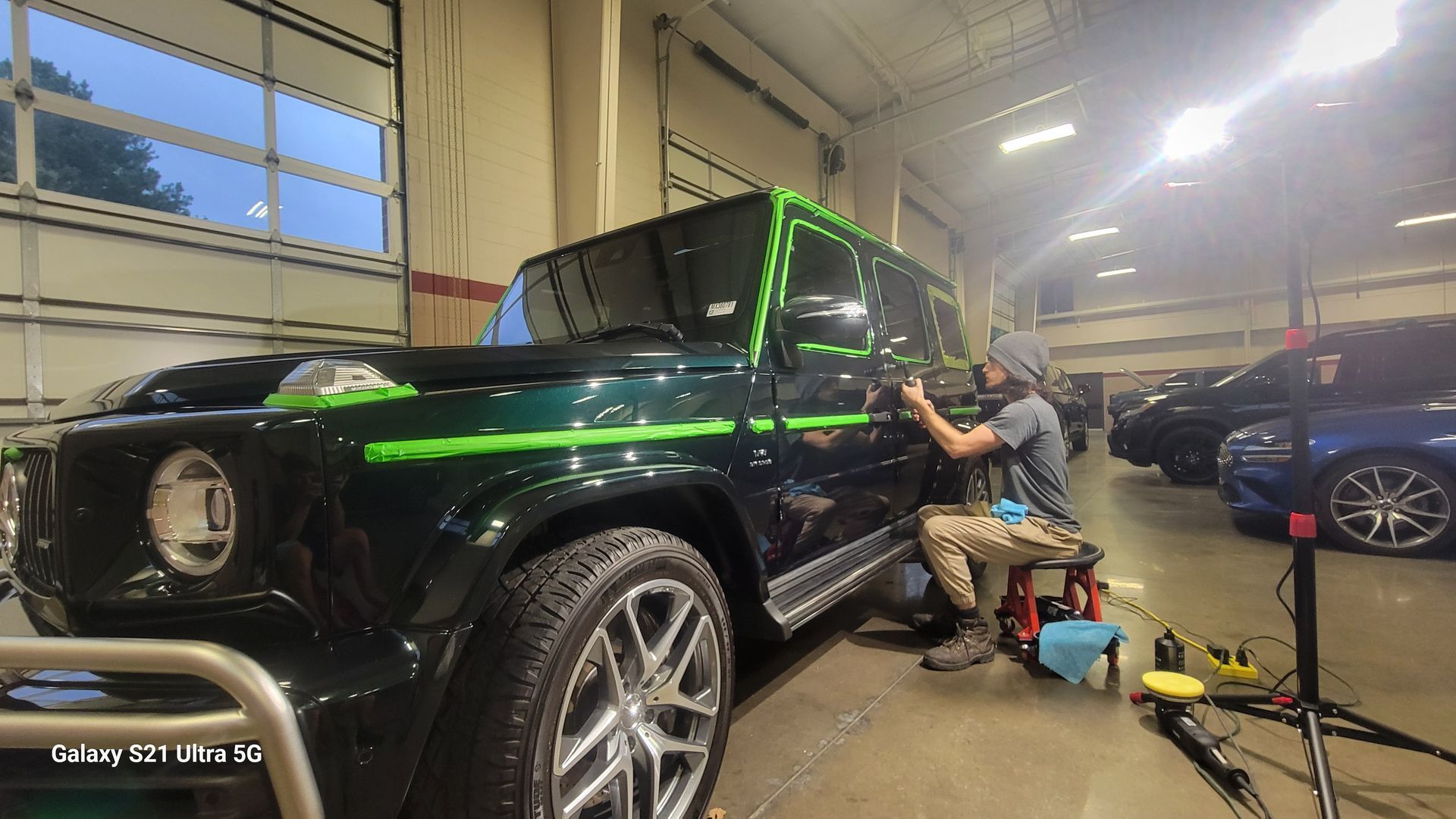 Black SUV in a garage while a person works on its side near tools and a blue car in the background