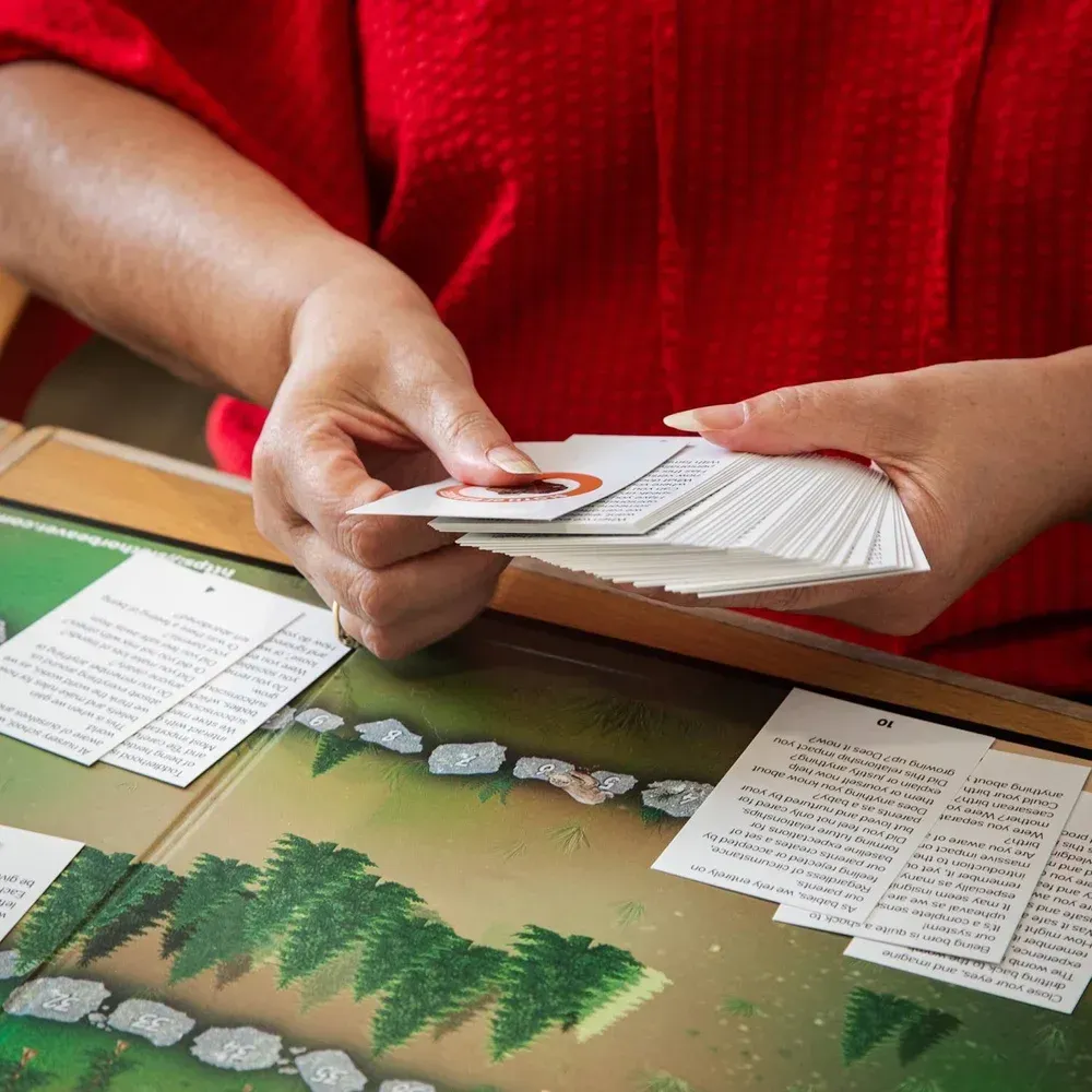 Hands hold a deck of playing cards over a board game with a forest-themed surface and printed cards.