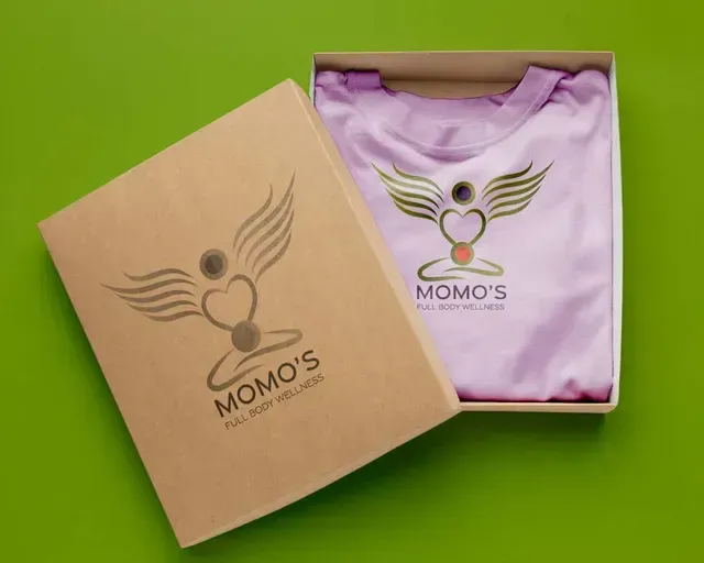 A light purple t-shirt in a tan gift box, both featuring a gold winged logo labeled 