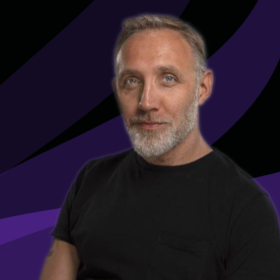 A man with a graying beard and blue eyes, wearing a black t-shirt, posed against a dark, abstract purple and black background.