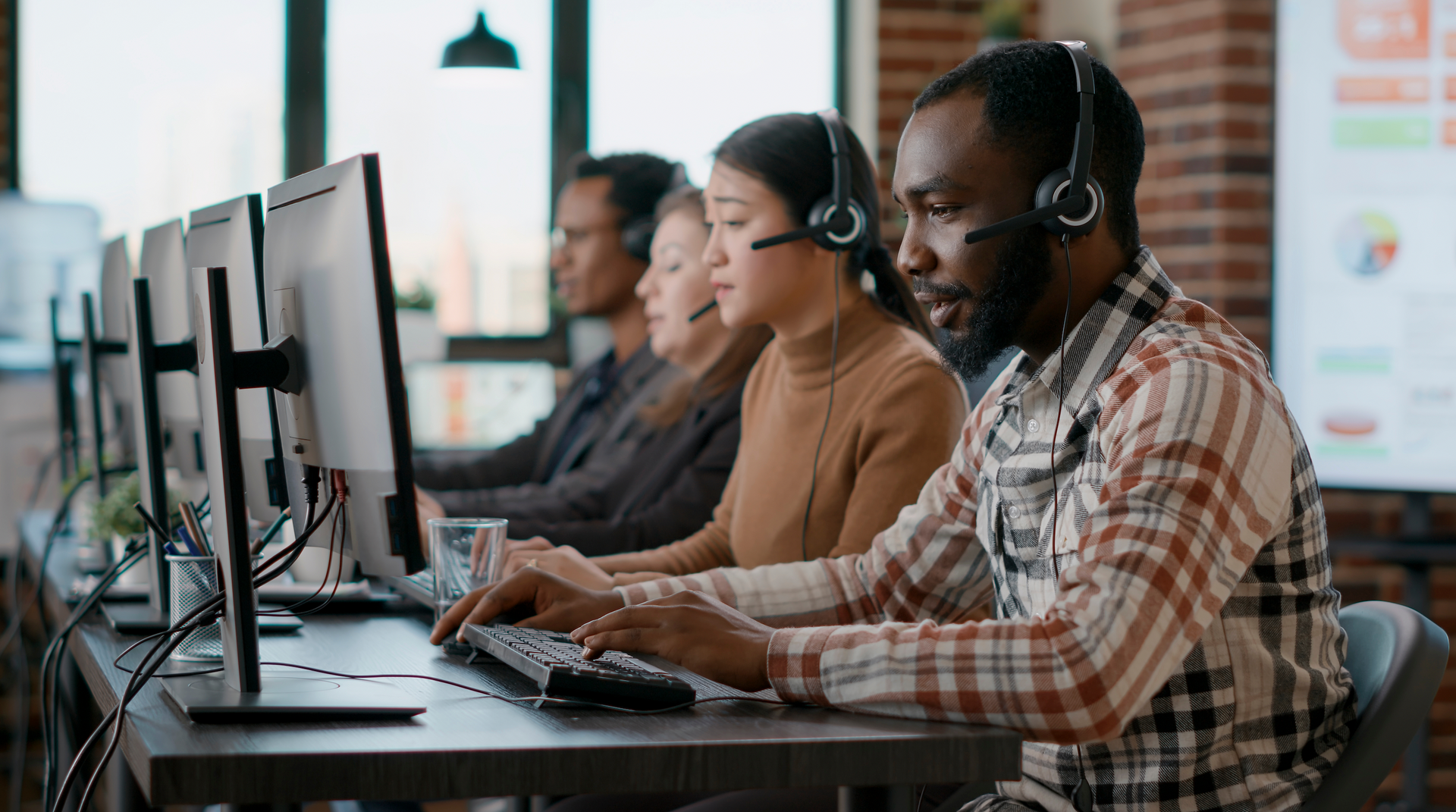 A diverse group of customer service representatives work at their computers wearing headsets. They are in an office setting.