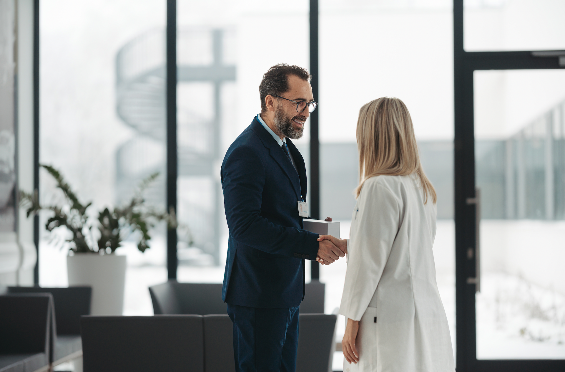 A man in a suit shakes hands with a woman in a white coat in a modern lobby. Both are smiling.