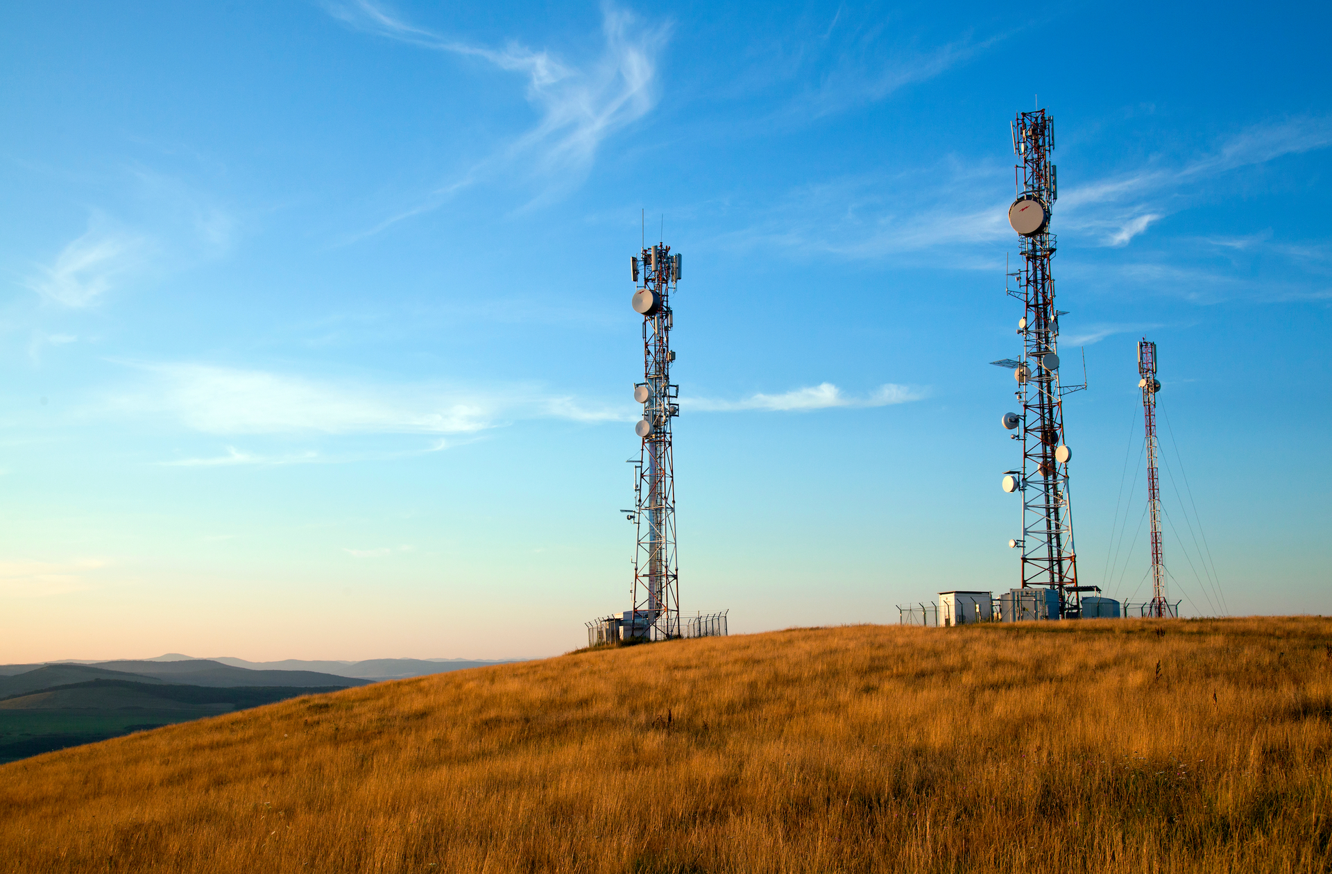 Three communication towers on a grassy hilltop against a blue sky.