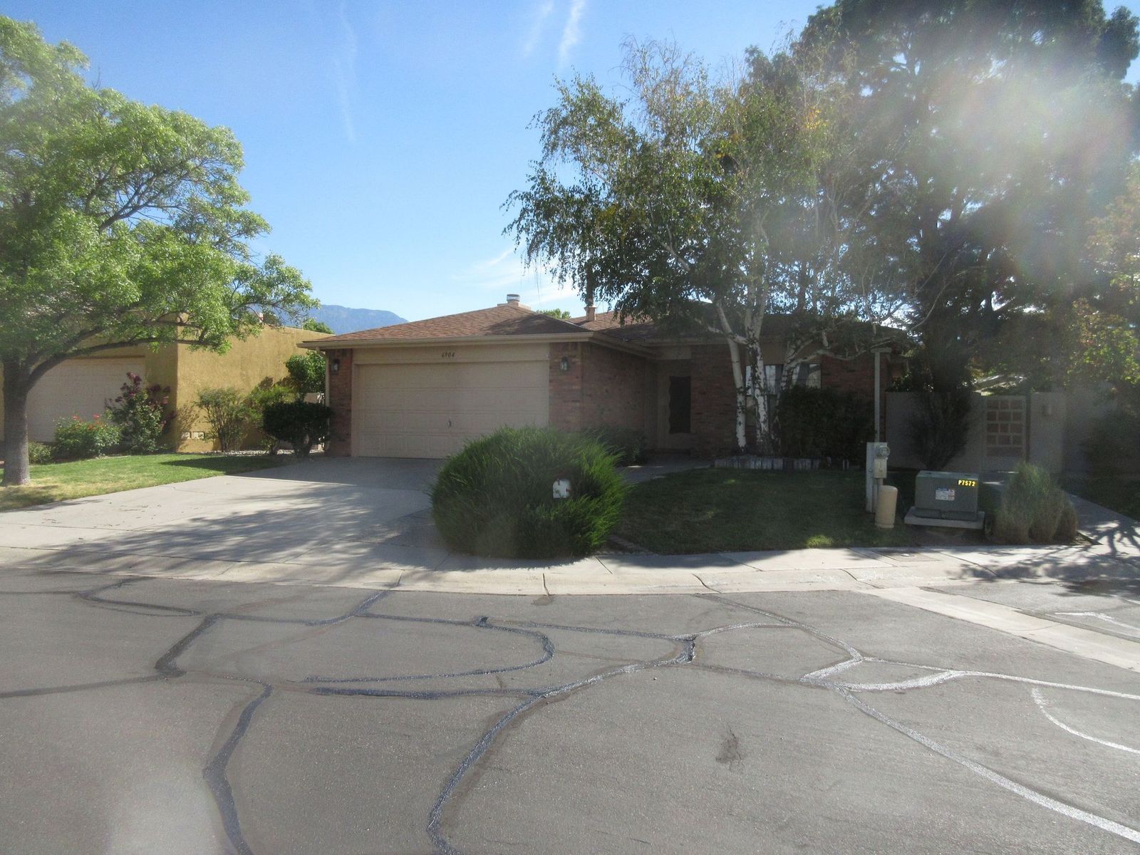 House with a two-car garage, set on a slightly sloped lawn, trees in front. Pavement in foreground with a blue sky.