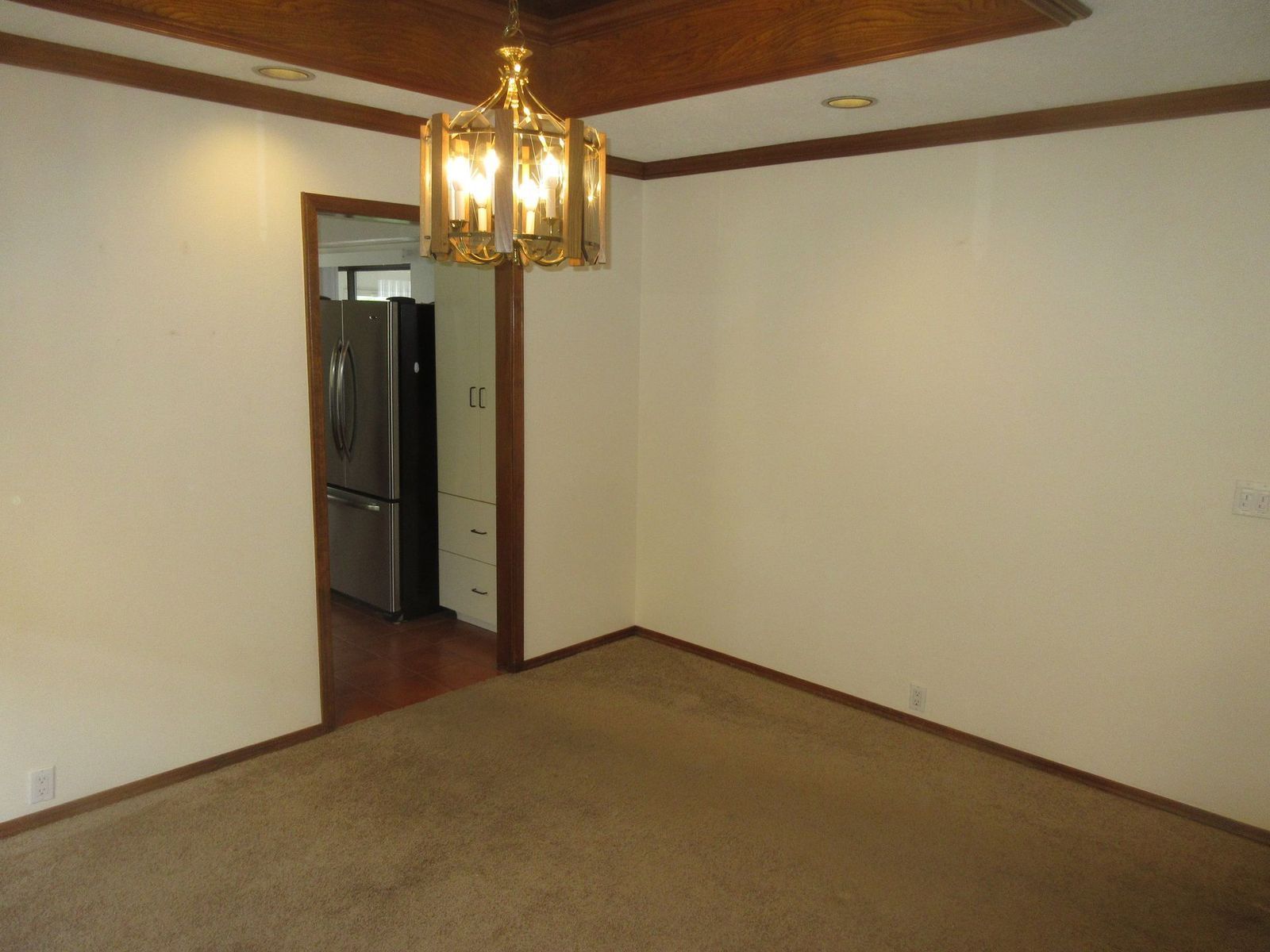 Empty room with beige walls, brown trim, and a gold chandelier; doorway leads to a kitchen.
