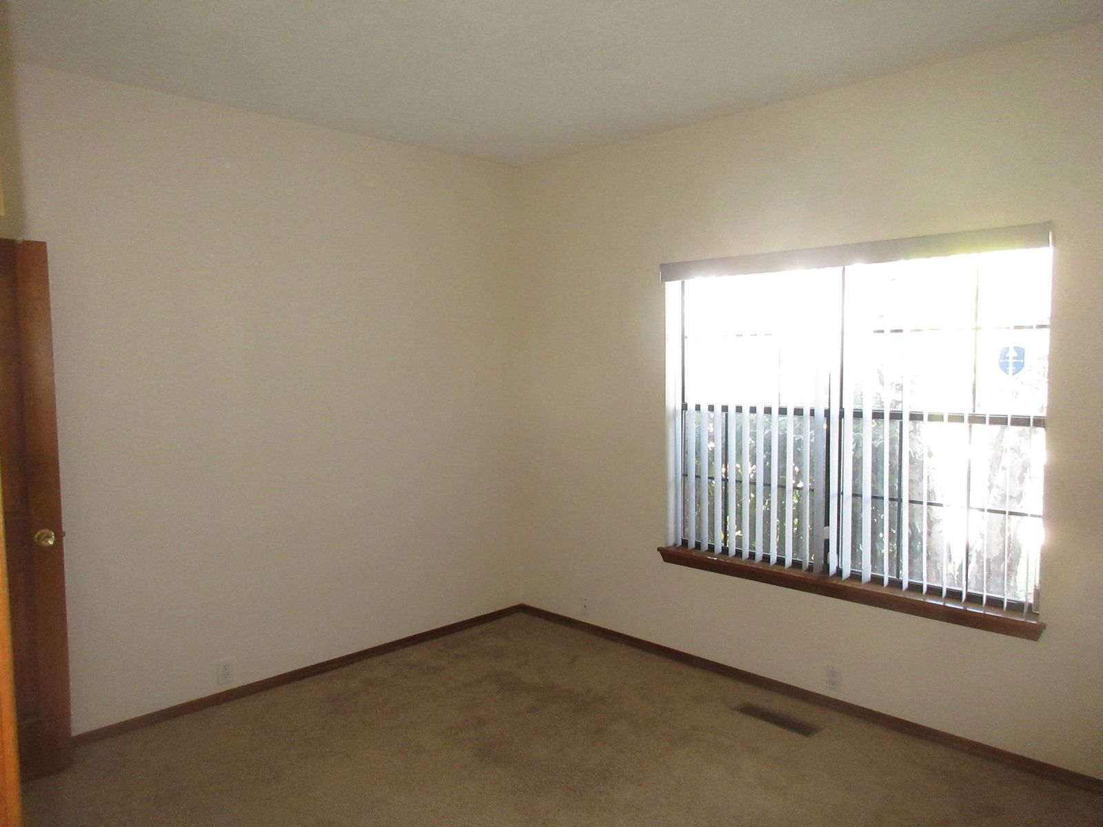 Empty room with beige walls, brown carpet, window with blinds, and a closed wooden door.