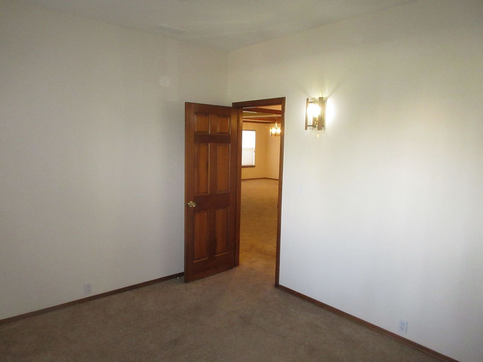 Empty room with doorway, brown door, beige walls, carpet, and sconce.