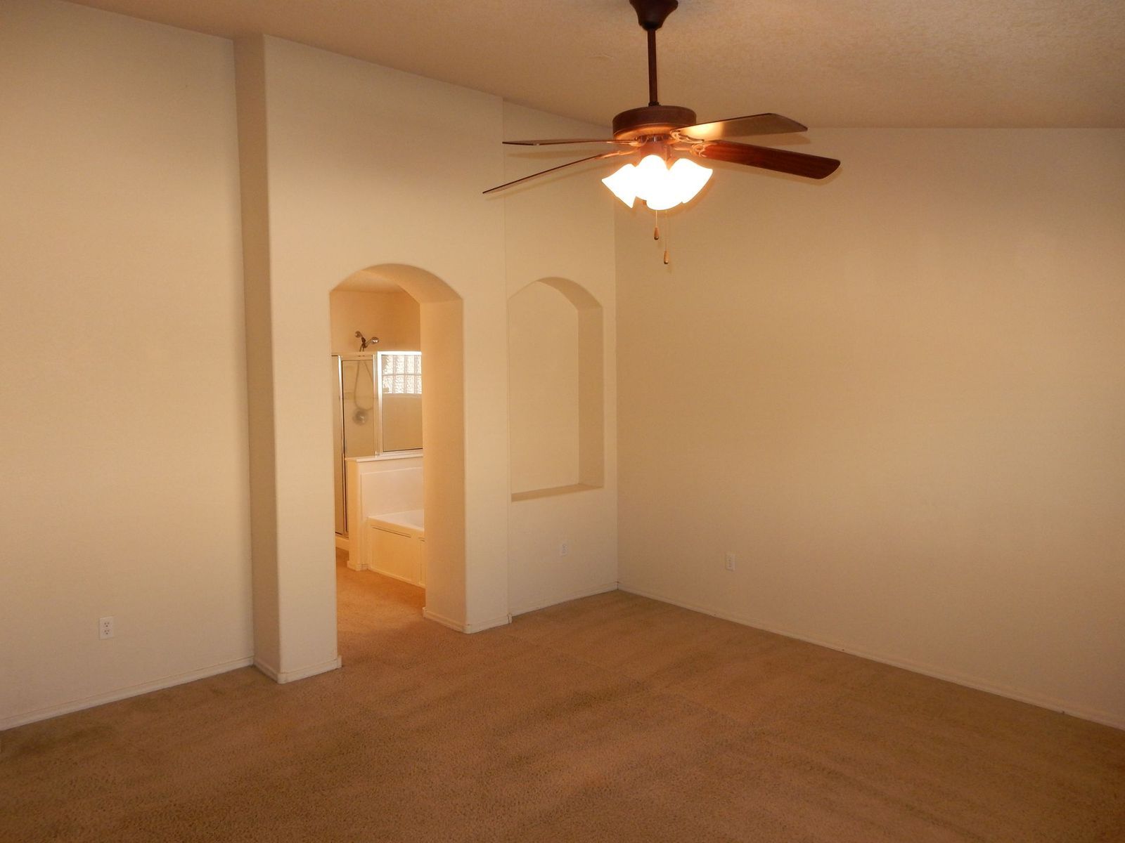 Empty beige-walled bedroom with arched openings. A ceiling fan hangs above brown carpet; a bathroom is visible.