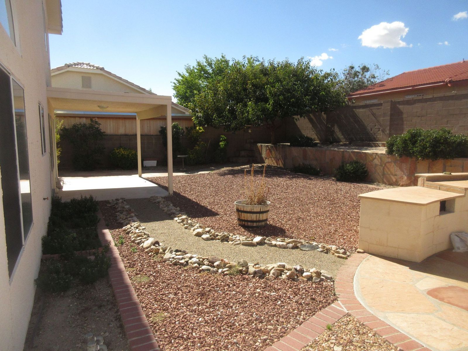 Backyard with red gravel, a brick-edged pathway, a patio cover, and a tree against a blue sky.