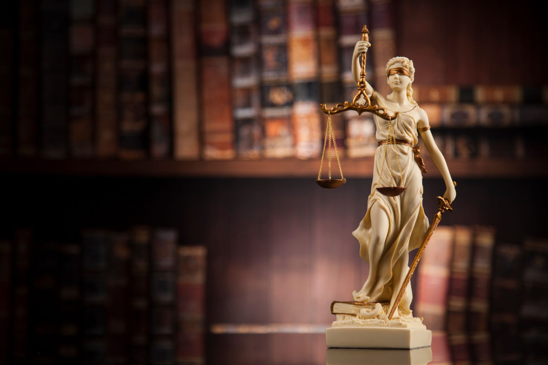 Statue of Lady Justice holding scales and sword, set in a library with bookshelves in background.