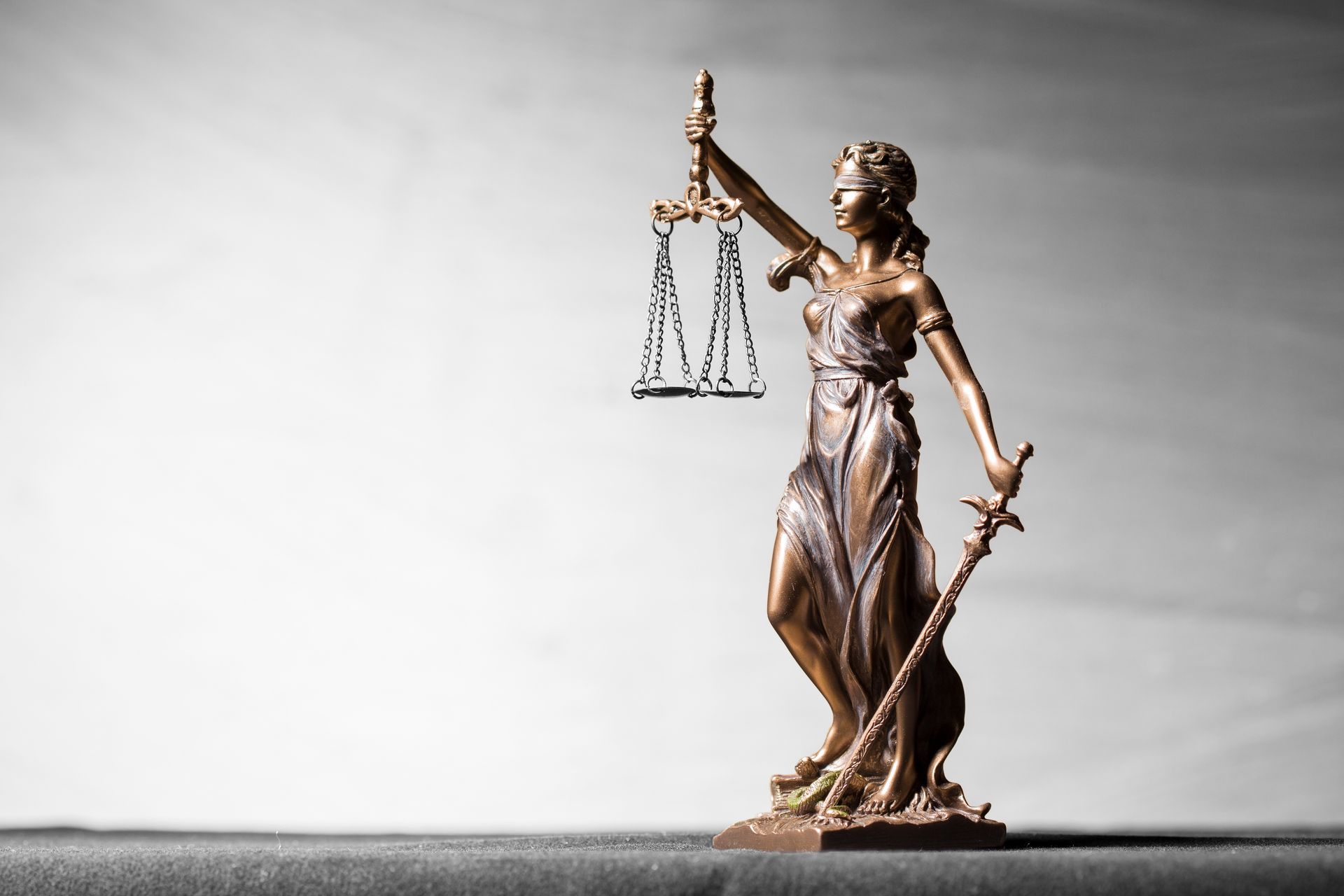 Statue of Lady Justice holding scales and sword. Bronze on a black surface against a gray background.