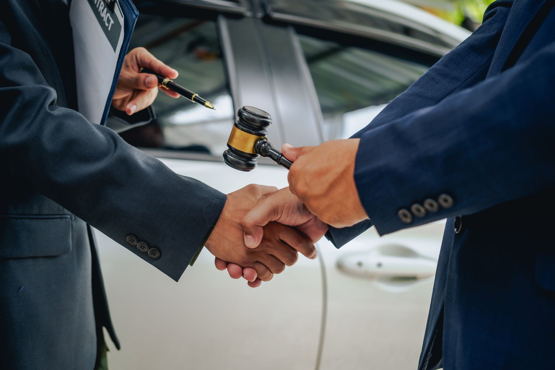 Two people in suits shaking hands near a car, one holding a gavel.