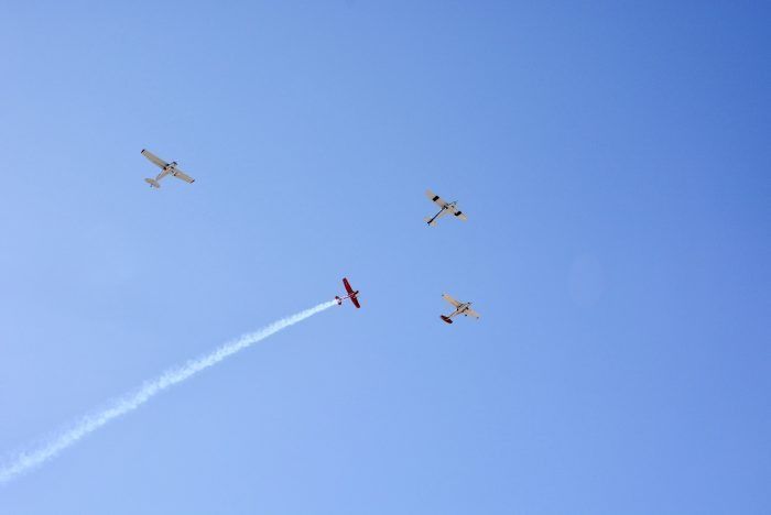 Three small planes in a blue sky, one leaving a white smoke trail while another flies red.