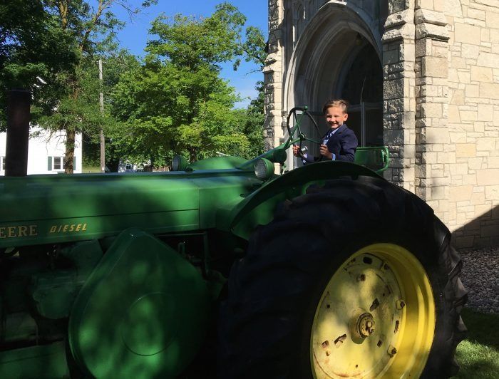 Child driving a green tractor beside a stone building in bright sunlight