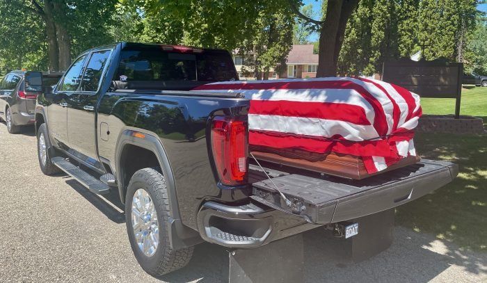 Black pickup truck carrying a U.S. flag-draped coffin in the bed, parked outdoors under trees