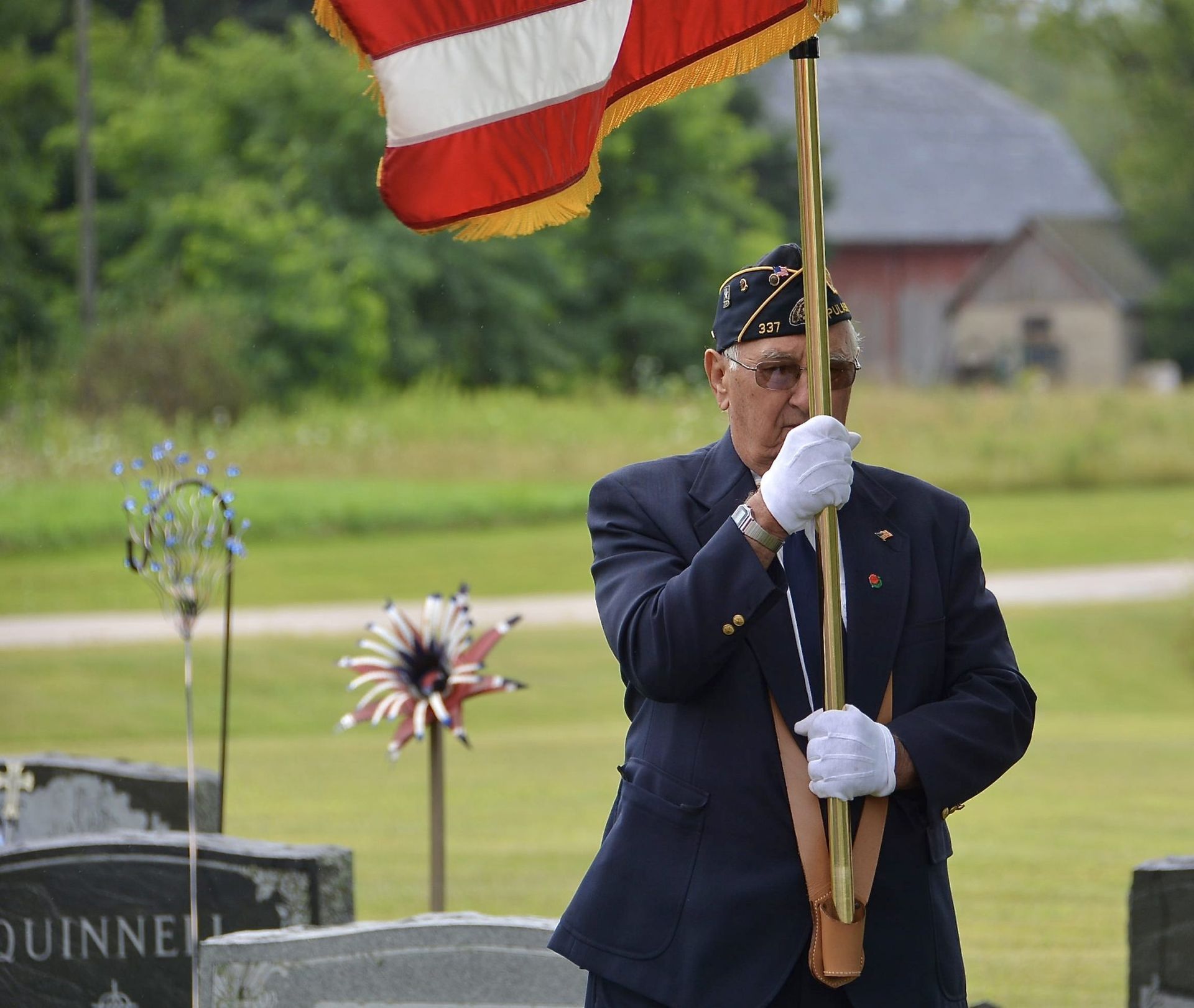 Veteran in dark uniform holding a U.S. flag at a cemetery memorial service