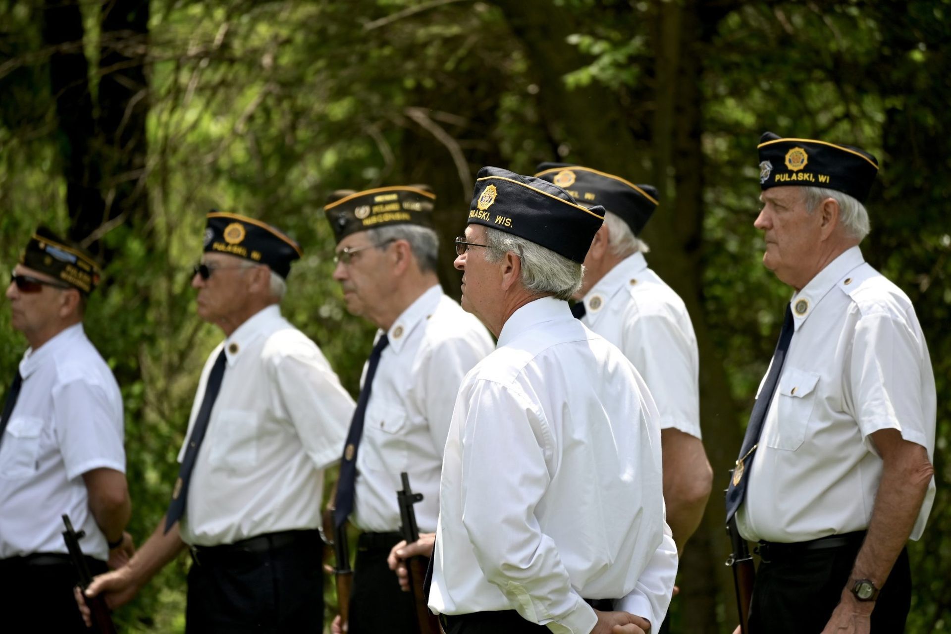 Veterans in white shirts and black caps standing in a row outdoors, side view