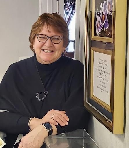 Smiling person in black sweater beside a framed plaque in a hallway