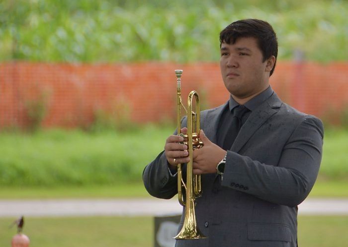Man in a dark suit holding a trumpet outdoors, with a blurred green and orange background