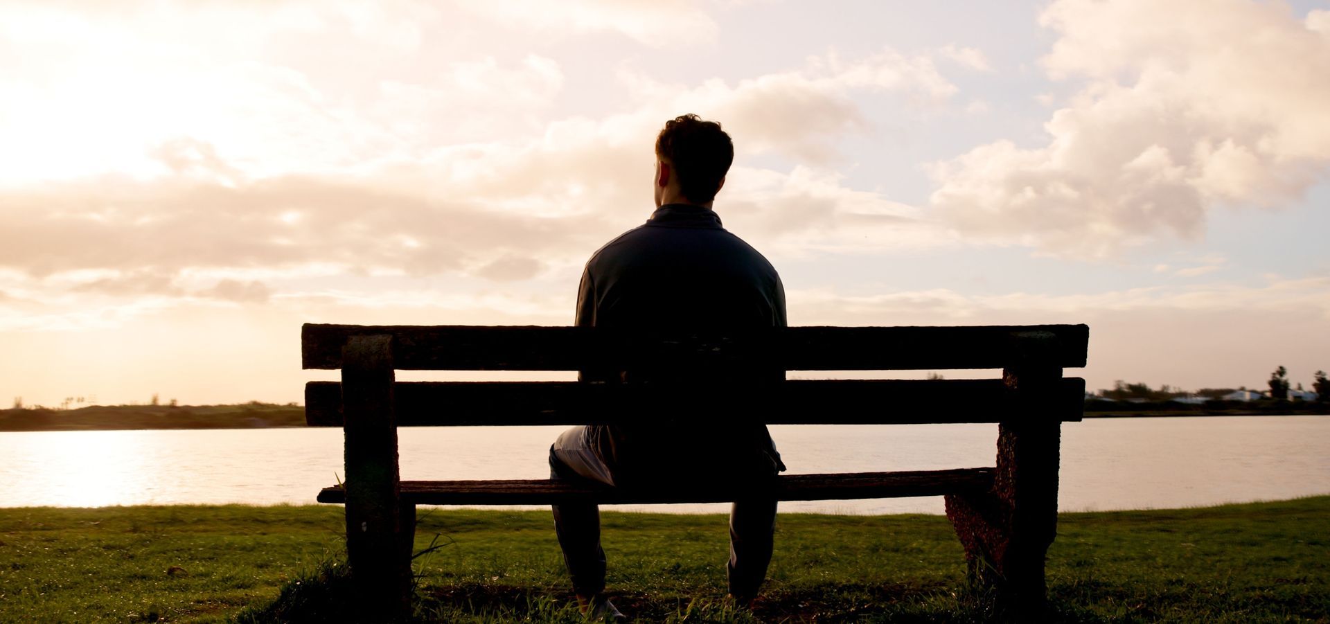 Person sitting on a bench by the water at sunset, facing the horizon.