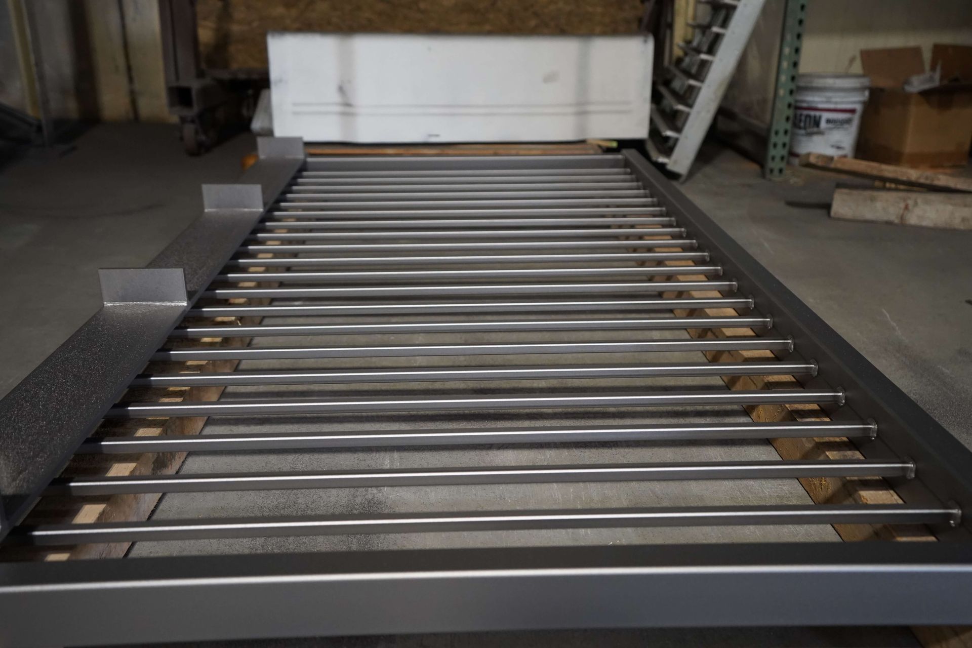 Metal gate with horizontal bars, partially constructed, on a wooden base in a workshop.