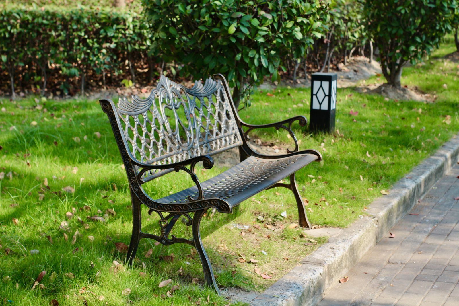 Ornate cast iron park bench with a dark bronze coated finish on green grass beside a brick walkway