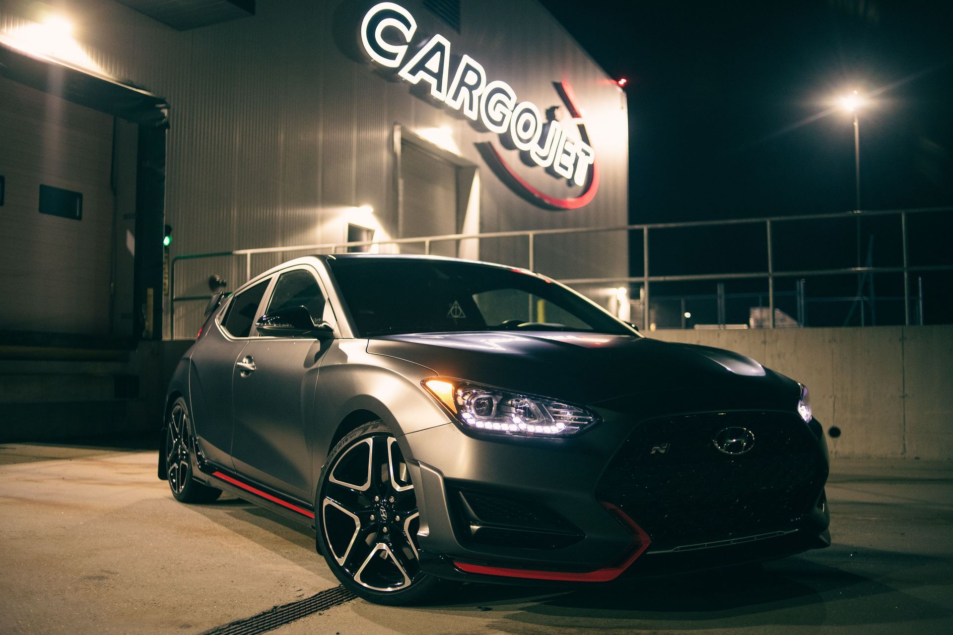 Dark gray sports car parked at night outside a lit garage with neon signage