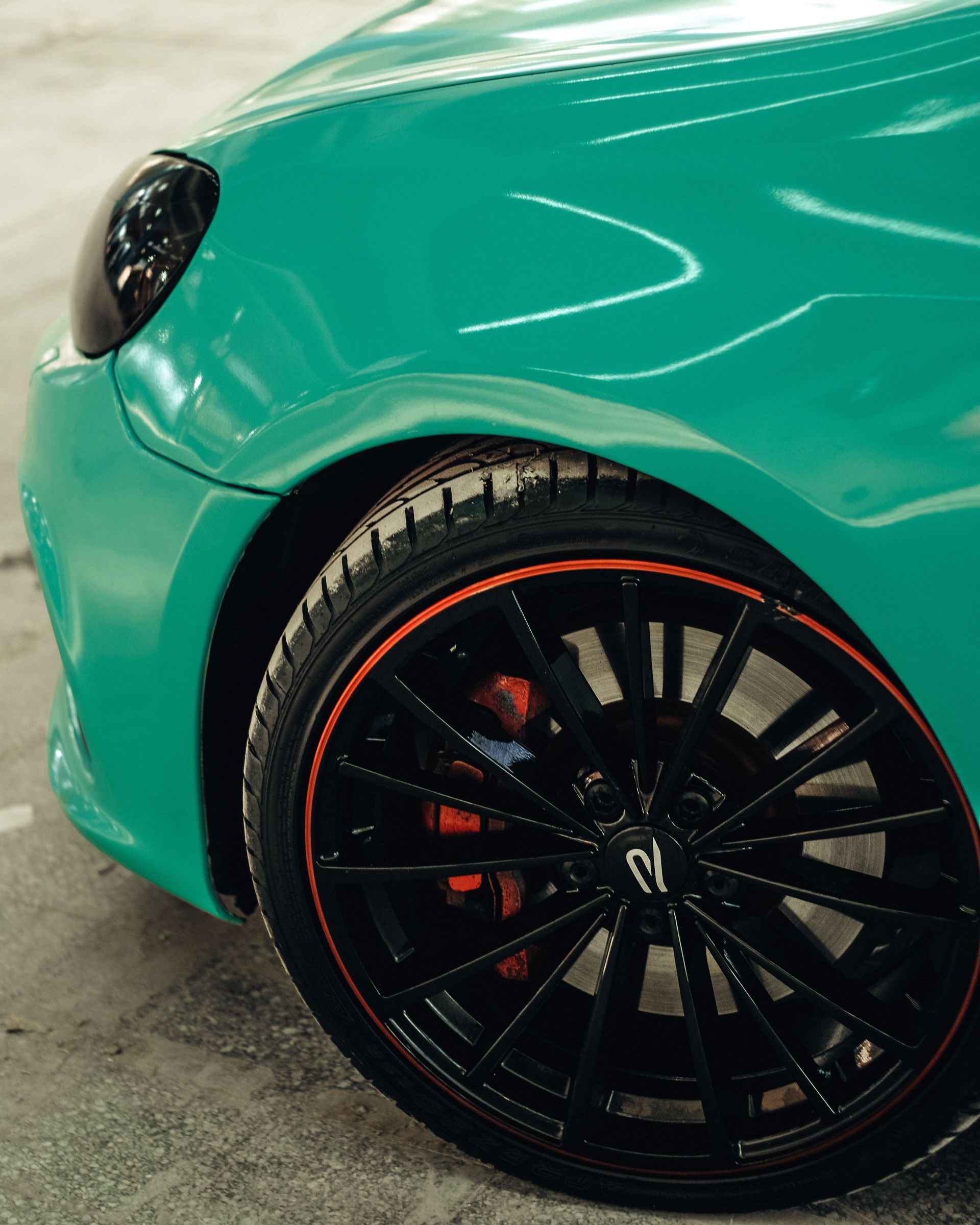 Close-up of a teal sports car’s front wheel with a black rim and orange accent line.