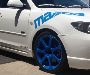 A white mazda with blue wheels is parked on the side of the road