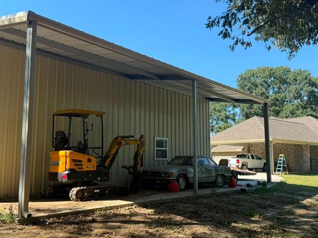 Yellow excavator under a metal carport beside parked vehicles and a house.