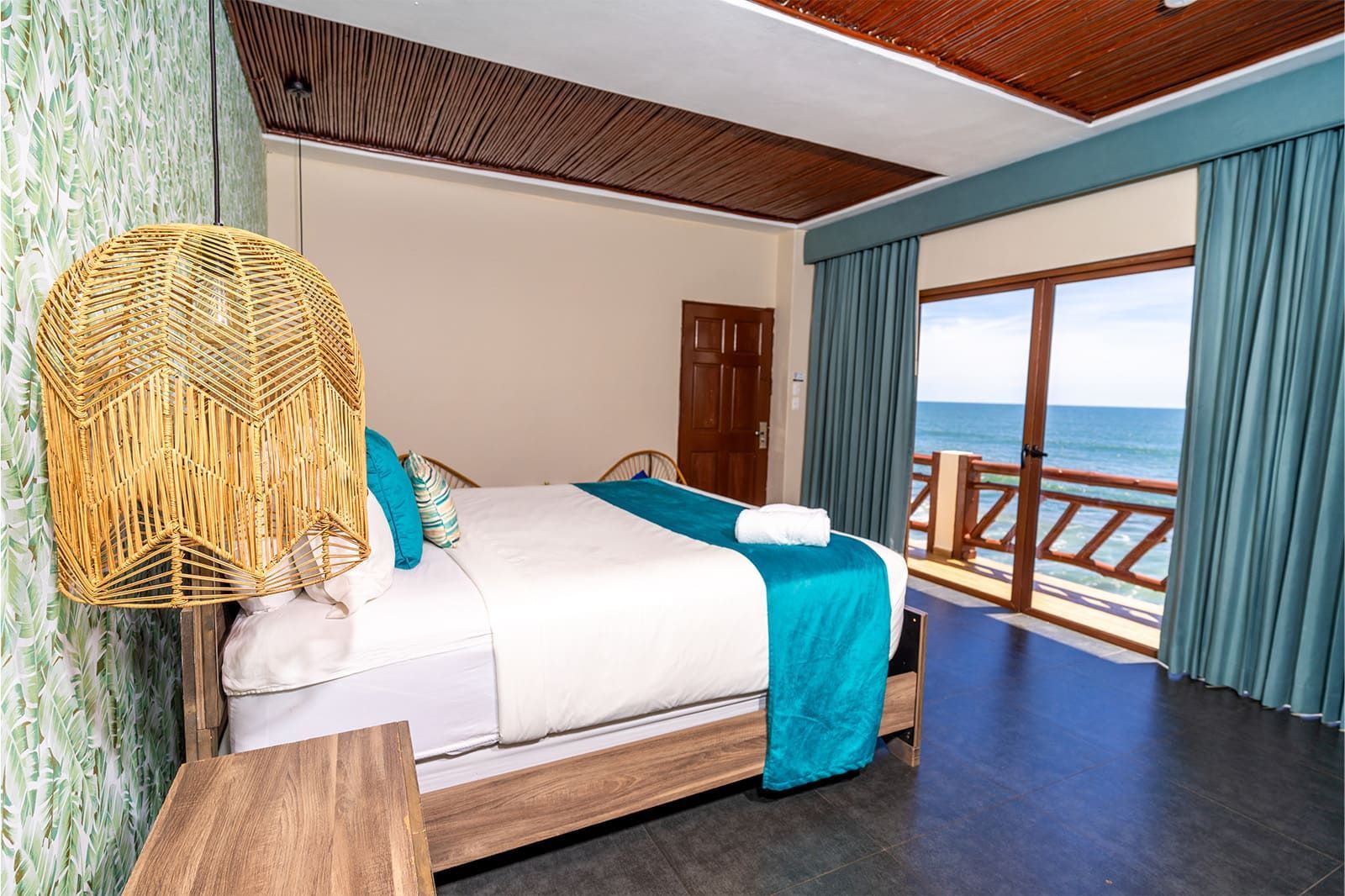 A hotel room with a queen bed featuring blue runners, a rattan pendant lamp, and a sliding door view of the ocean.