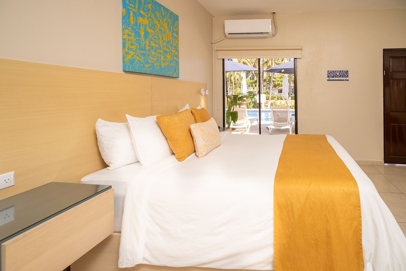 A hotel room with a large bed, white linens, yellow accents, a blue wall painting, and a sliding glass door to a patio.