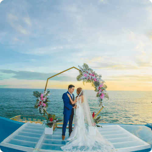 A couple standing together under a floral-decorated hexagonal arch on a platform overlooking the ocean at sunset.