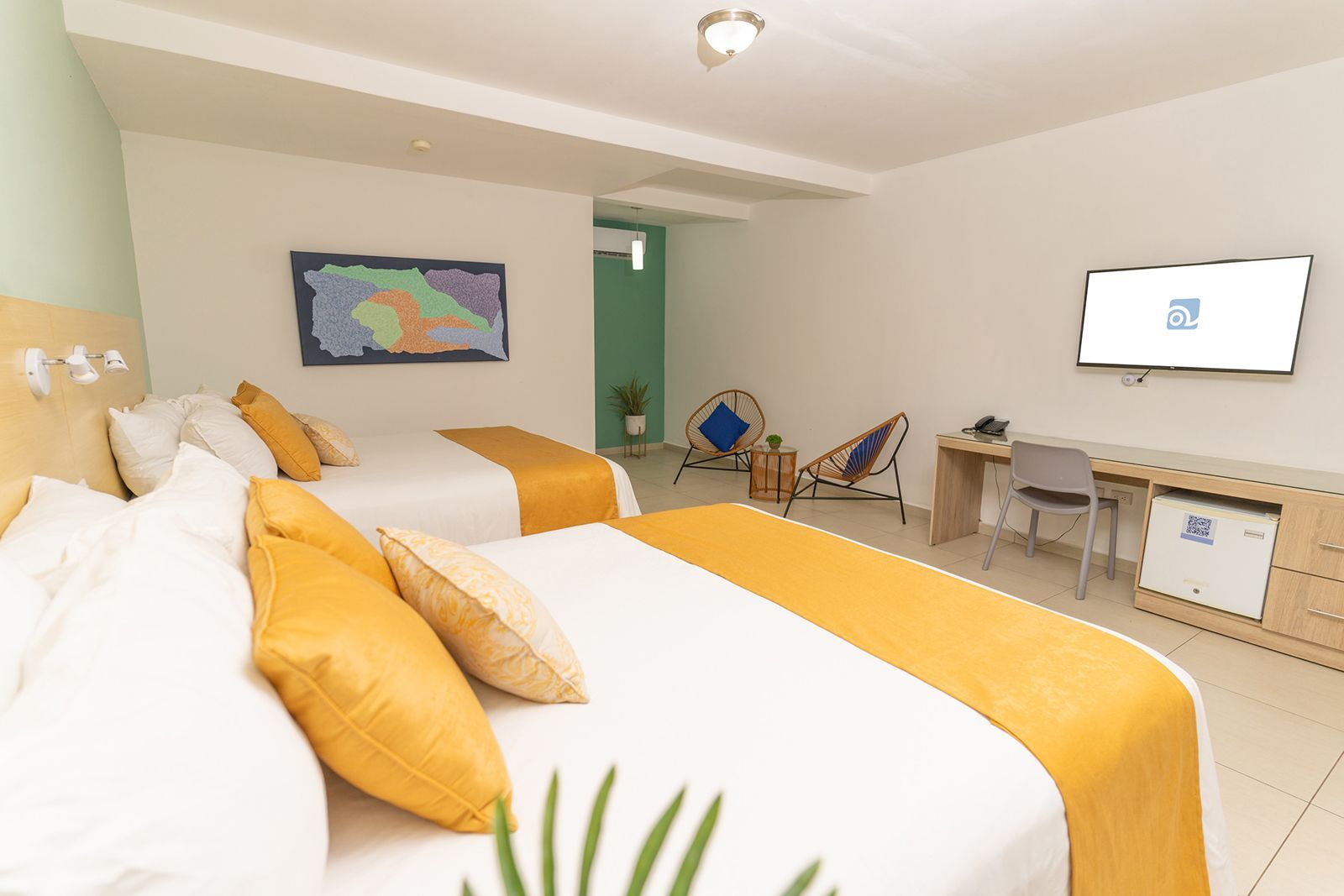 A hotel room with two white beds, yellow runners and pillows, a desk, a television, and two rattan chairs.