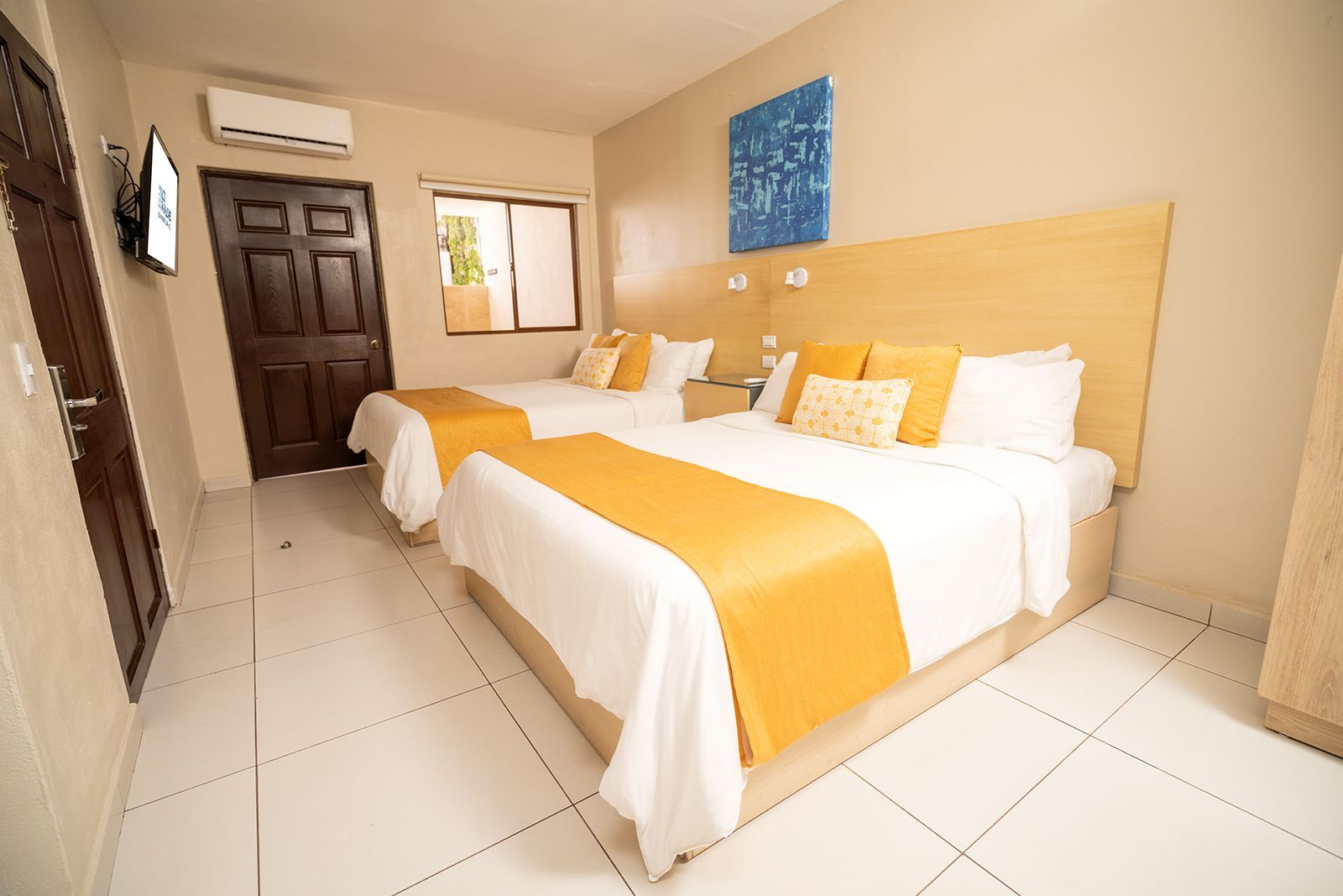 A hotel room with two white beds featuring yellow runners, wooden headboards, white tile floors, and a dark brown door.