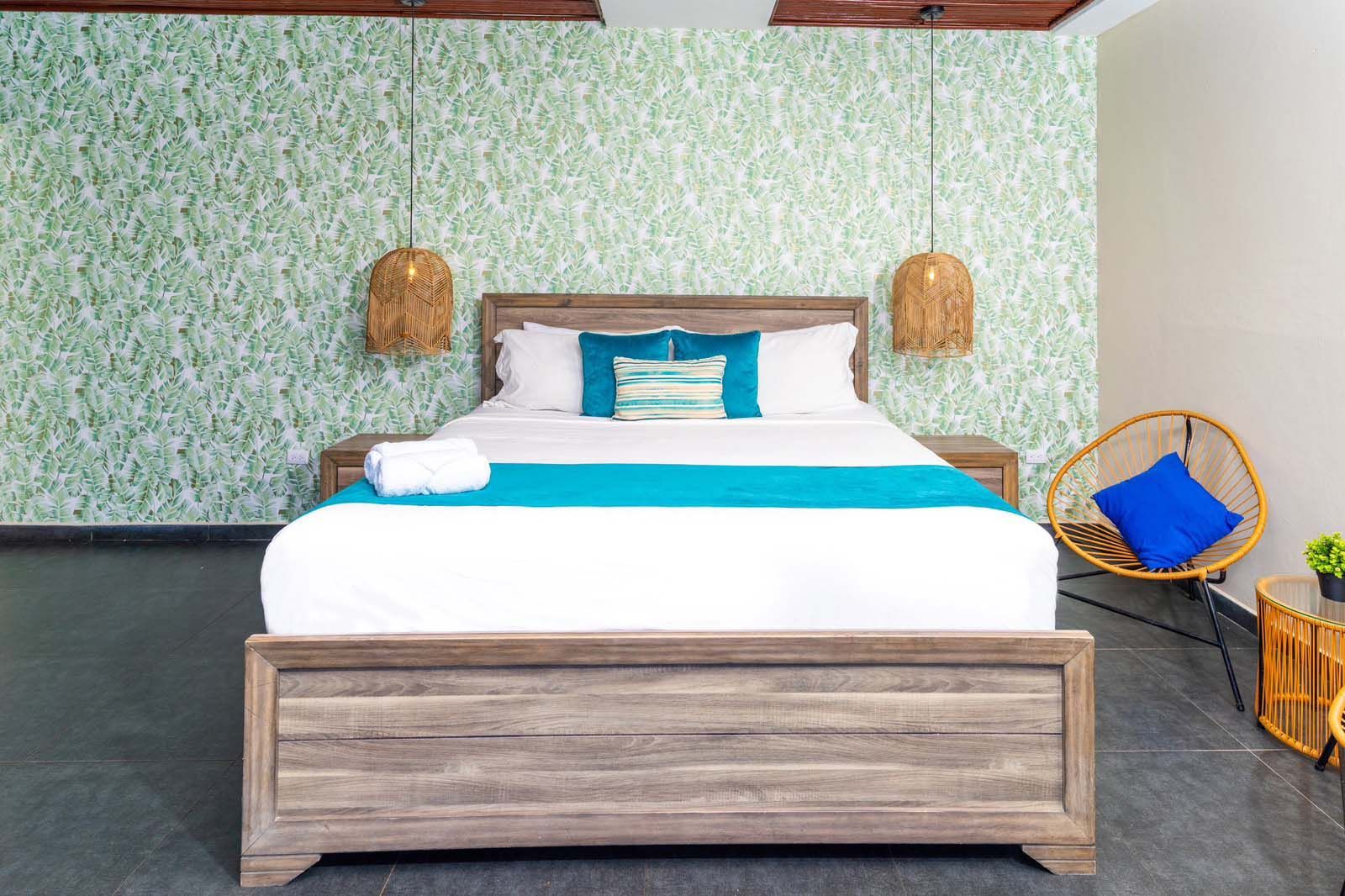 A hotel room with a wooden bed, patterned green wallpaper, two hanging wicker pendant lights, and a blue accent chair.