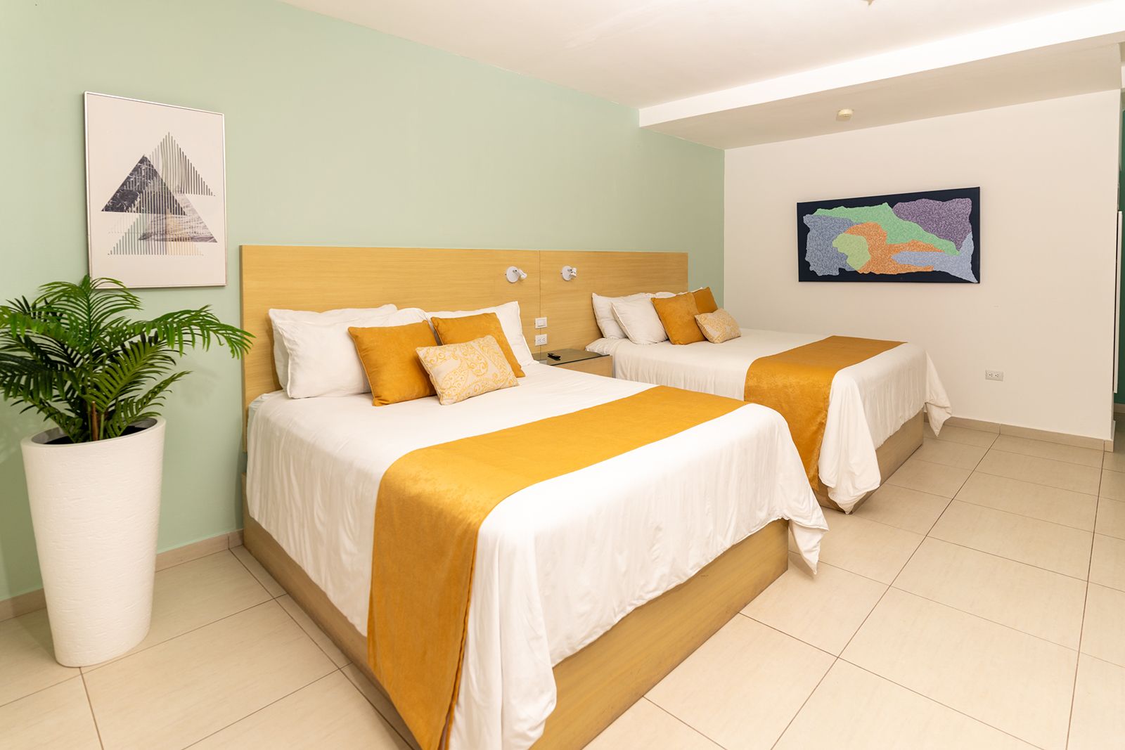 A modern hotel room with two queen beds, pale green walls, a potted plant, and framed artwork on white tile flooring.