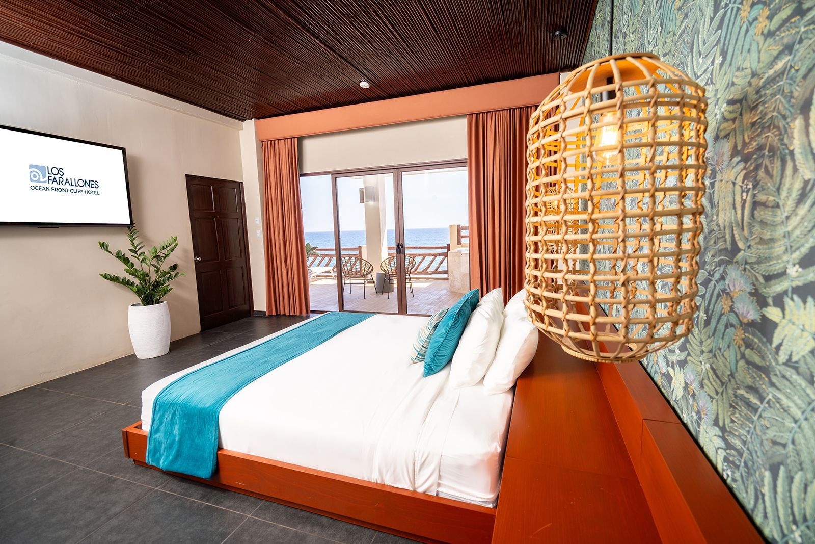 Modern hotel room with a bed, teal accents, wicker pendant light, and ocean view through open doors
