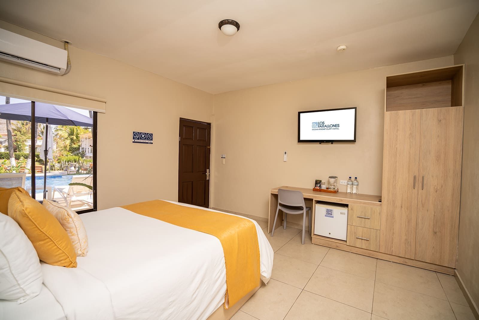 A brightly lit hotel room featuring a queen bed with yellow accents, a desk with a TV, a mini-fridge, and a wooden wardrobe.