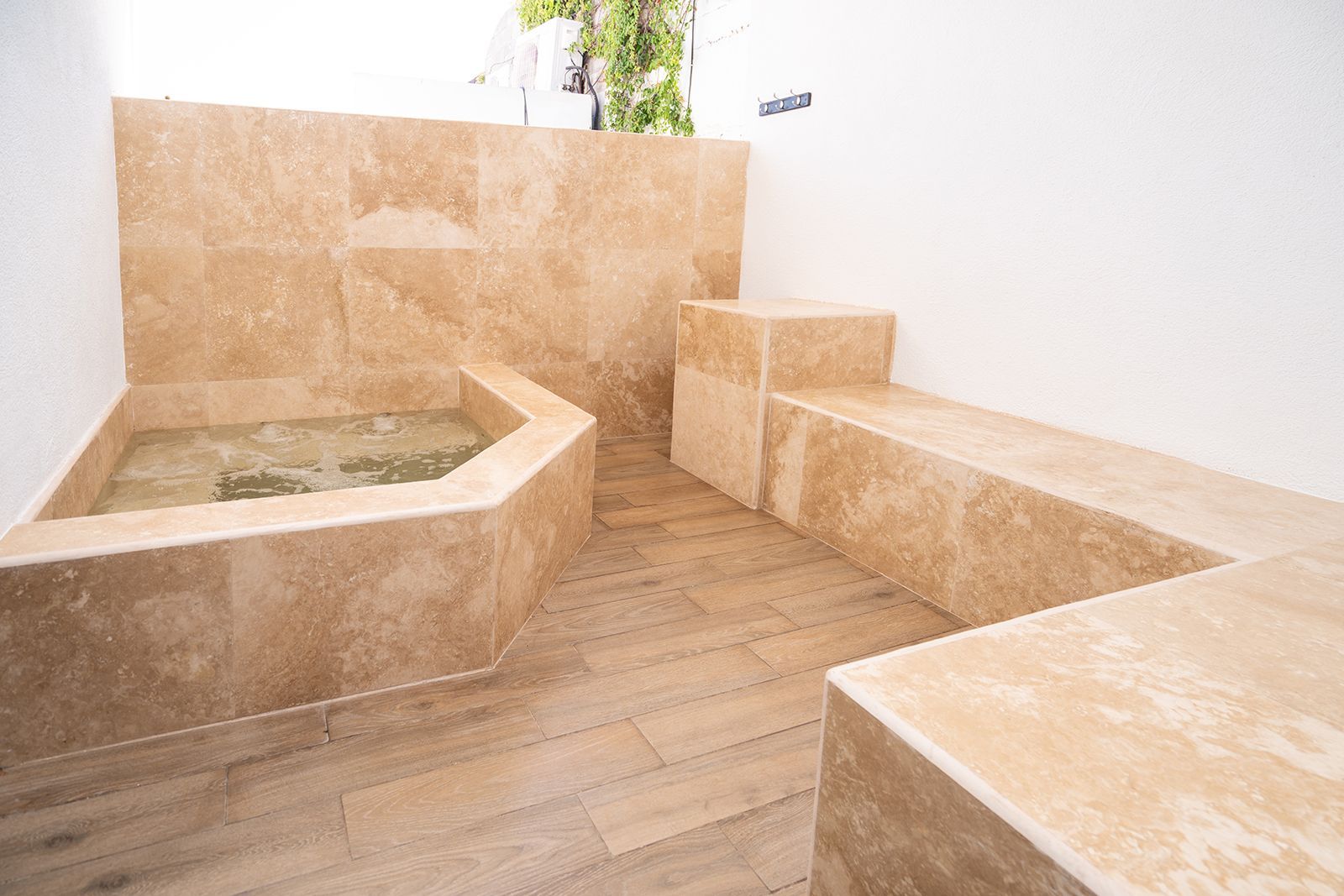 A small outdoor stone pool next to a built-in stone bench, both finished in tan travertine tiles on a wood-look floor.