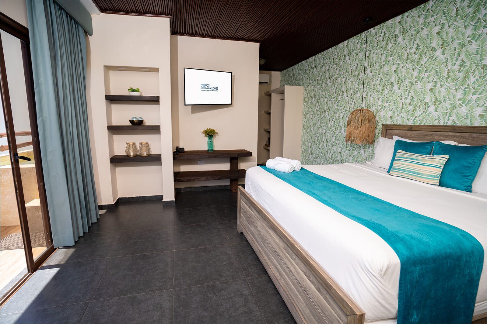 A hotel bedroom with a large bed, teal accent runner, tropical-patterned wallpaper, and a wall-mounted TV.