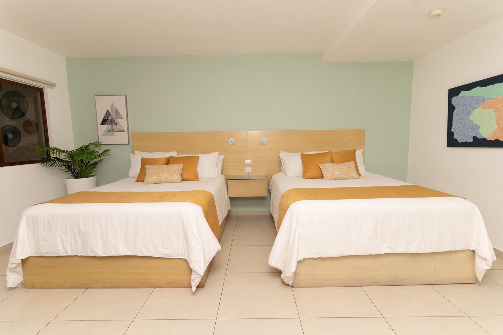 A hotel room with two queen beds featuring white bedding, tan runners, light wood headboards, and soft green walls.