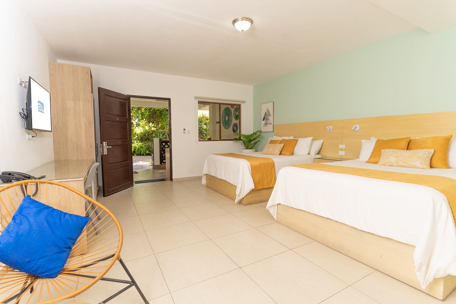 A bright hotel room with two queen beds, a rattan chair with a blue pillow, a desk, and an open door to a green garden.